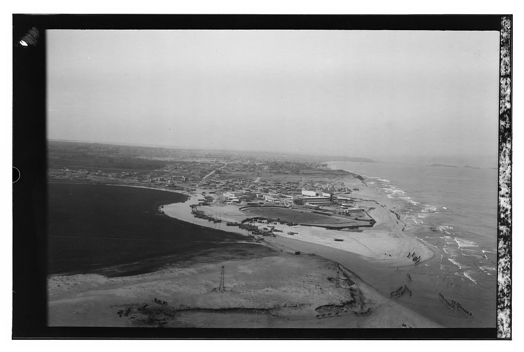 Black and white aerial photograph of Tel Aviv, a dense grid of flat rooftops running to the shoreline.